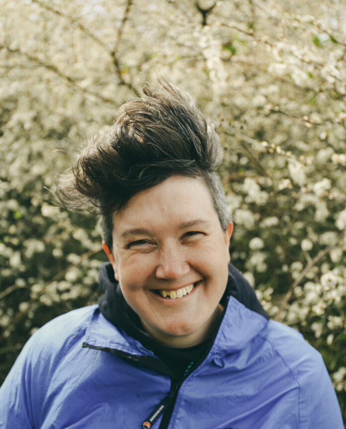 white woman with short brown hair, smiling and standing outside in front of a bush.