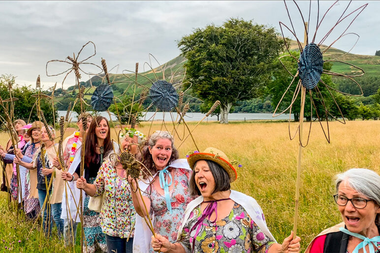 A group of women singers in floral costumes and with hand-held willow sculpture singing in a wildflower meadow with a lake and maountains in the distance
