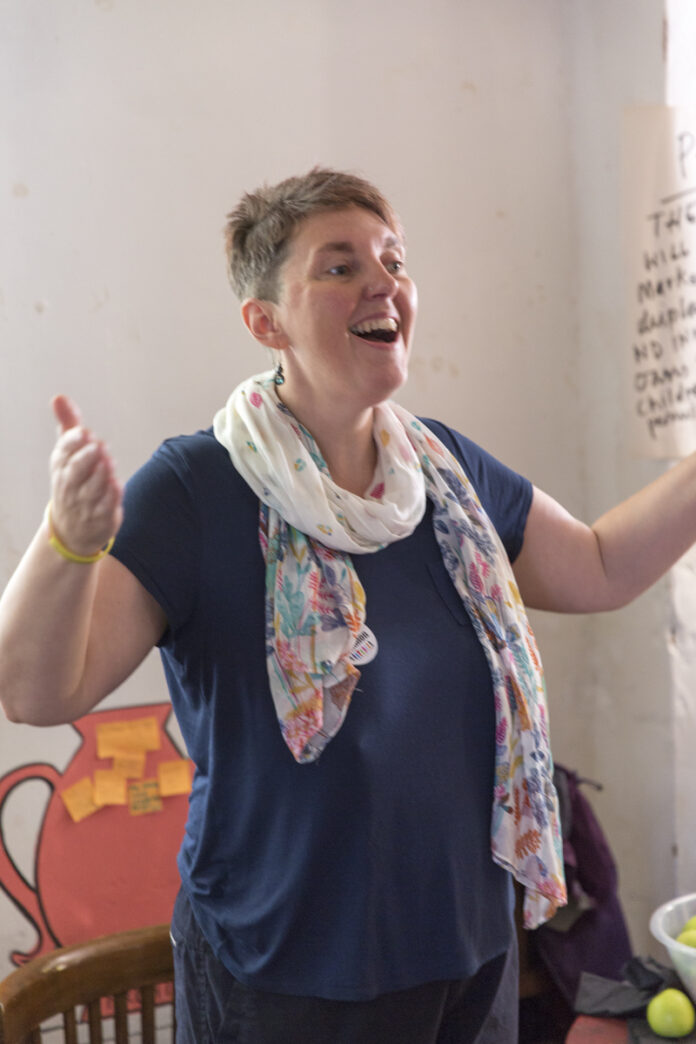 woman wearing blue top and white scarf, with arms wide and mouth open singing