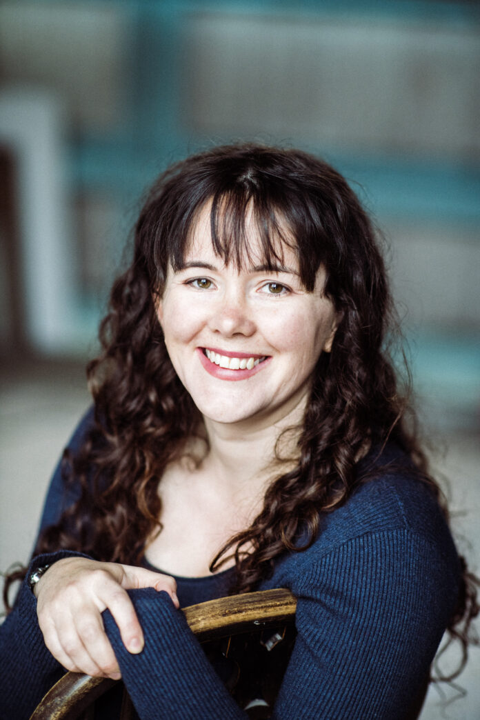 A photo of Daisy. She is sat on a chair smiling at the camera. She is a white woman with curly brown hair and is wearing a blue top.